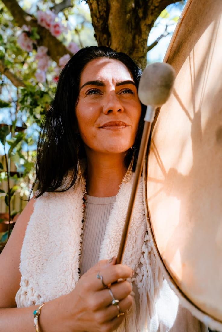Kati holding a drum beater beside her drum, smiling toward the light