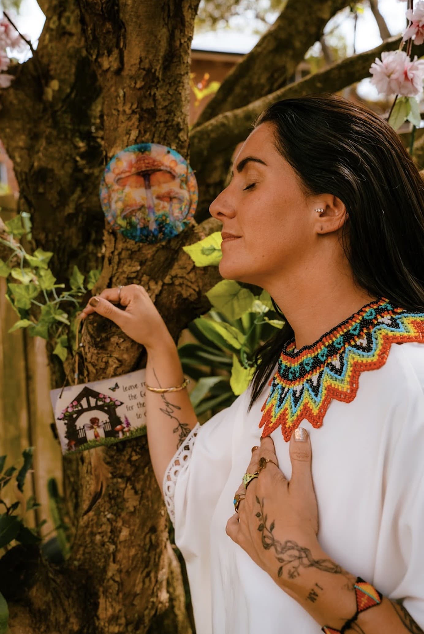 Kati by a tree in a beaded collar, eyes closed, one hand on her heart