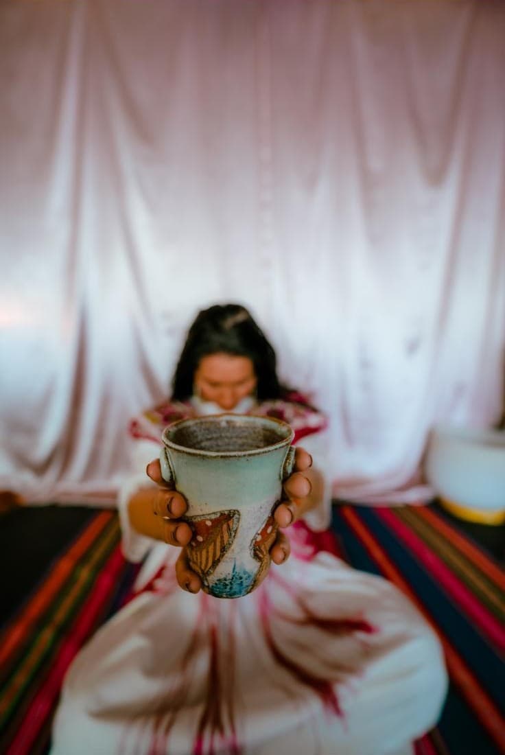 A woman seated on a woven rug, holding a cup in both hands