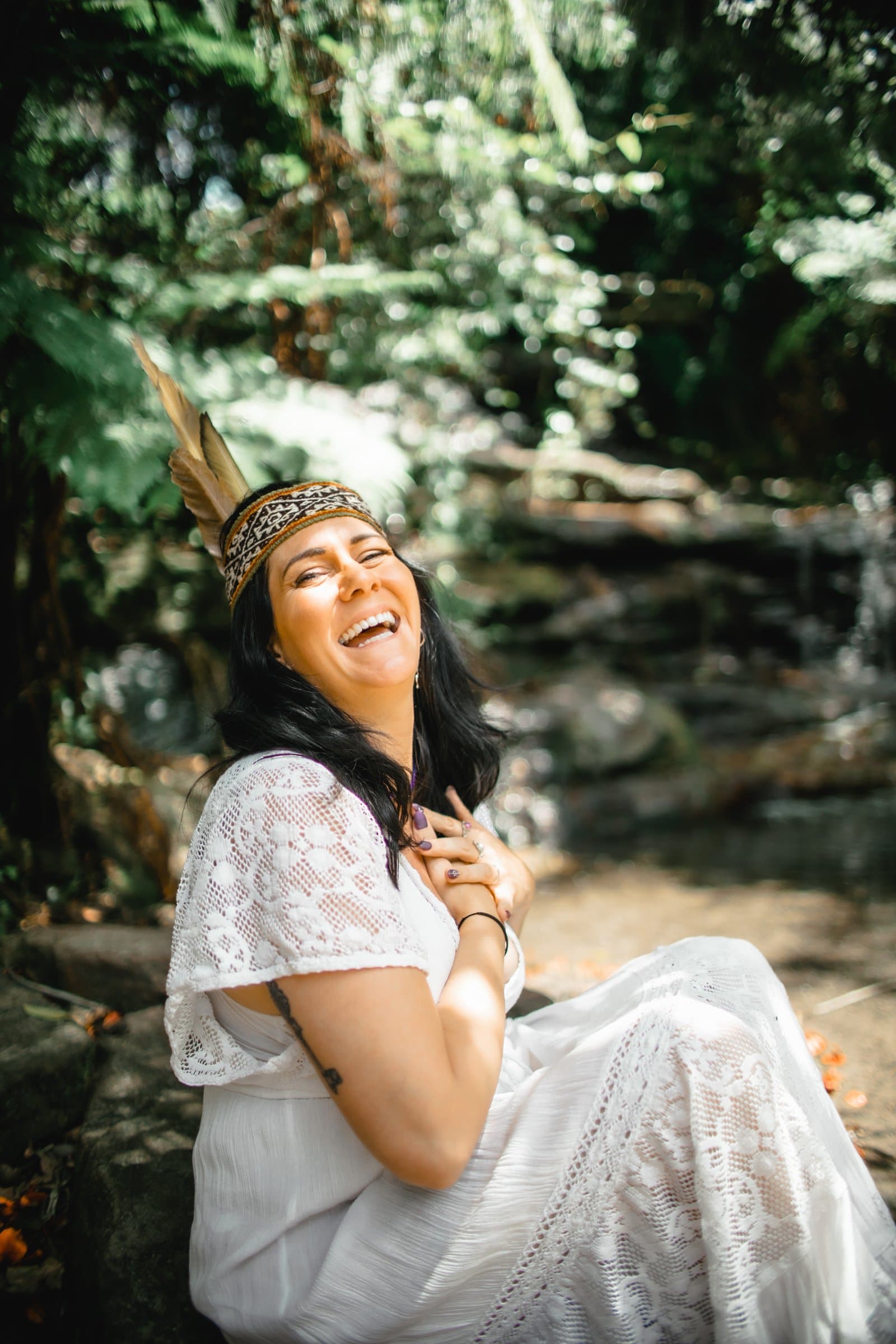 Kati laughing, hands on her heart, in a white lace dress and feathered headband by a forest stream