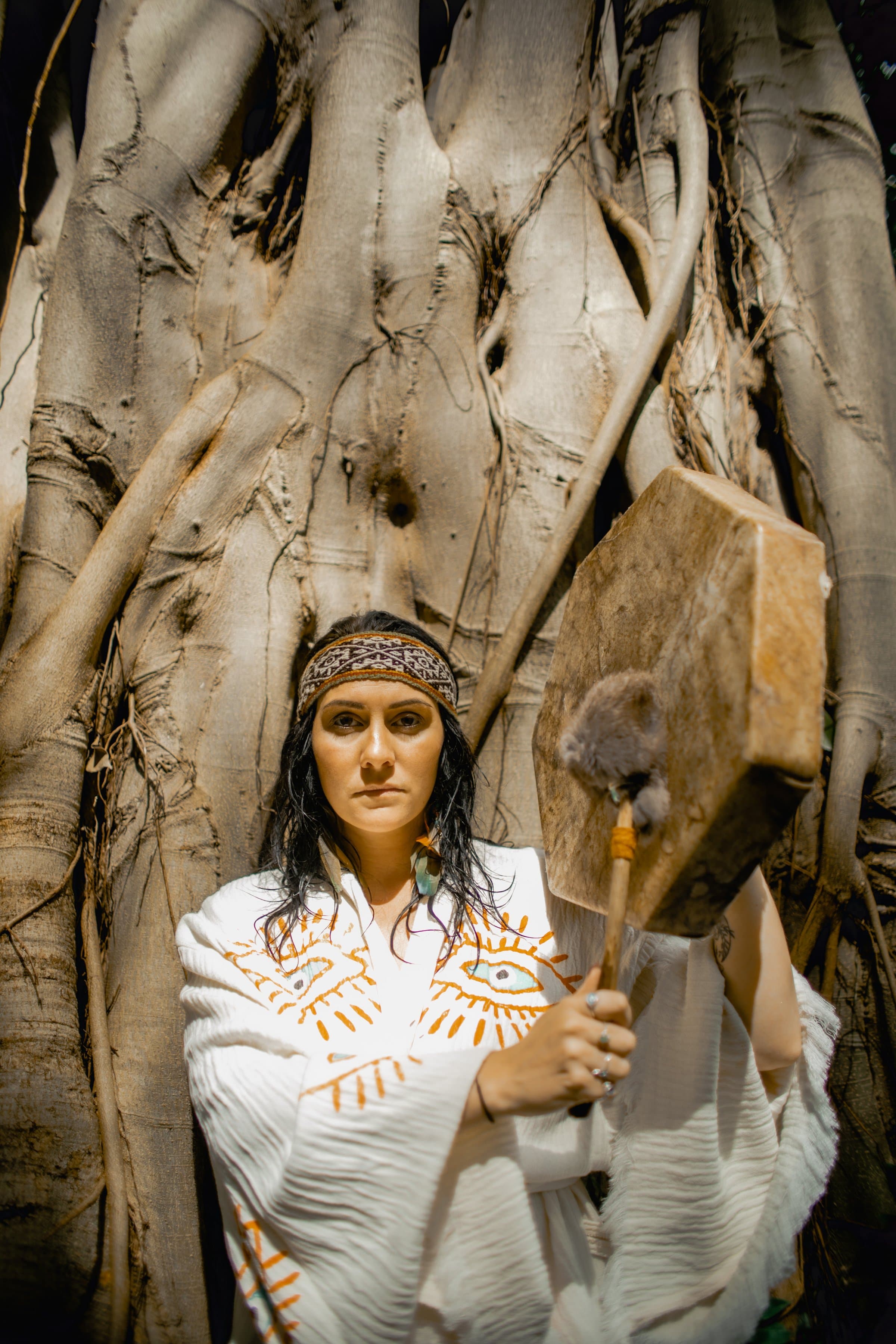 Kati in a woven headband with her drum, among the roots of a fig tree